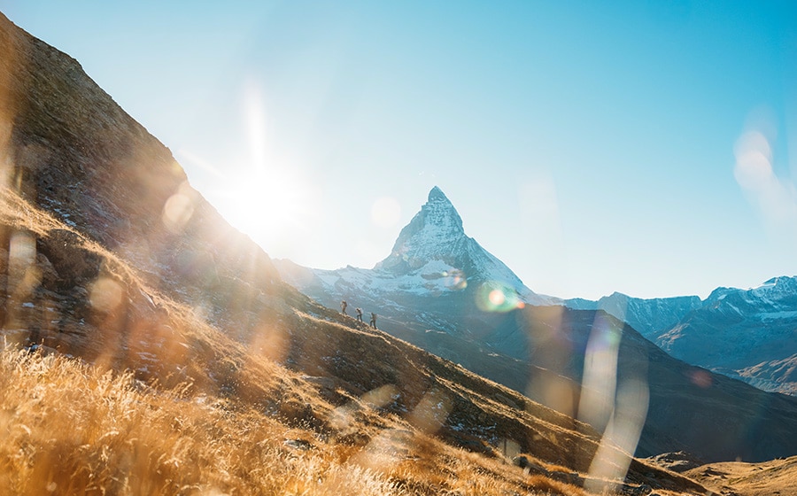 Photo of a mountain landscape featuring a striking, snow-capped peak beneath a bright blue sky and in brilliant sunshine. Golden grass in the foreground and two small hikers on a mountain ridge emphasize the vastness of the landscape and convey a sense of adventure in the great outdoors.