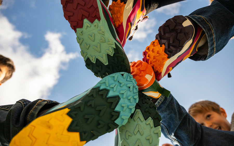 The photograph shows several children bringing their colorful LOWA shoes with different sole patterns together in the center of the frame. The low-angle shot emphasizes diversity and dynamism through bold colors and a contrasting sky in the background.