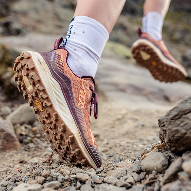 Close-up of a trail running shoe on a grassy path, with tattooed legs.